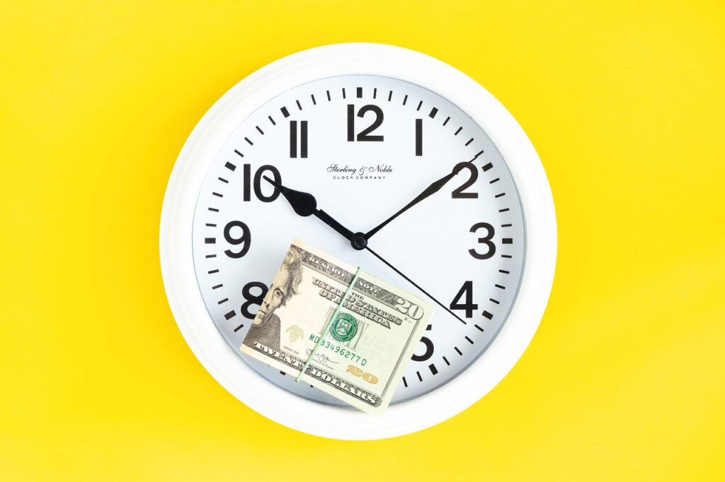 시계 위에 놓인 달러 지폐 이미지로, 시간이 지남에 따라 돈의 가치가 변하는 인플레이션과 투자 수익의 관계를 상징하는 사진. Image of a clock with a dollar bill symbolizing the time value of money and the impact of inflation on long-term investment returns.